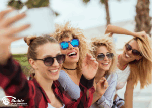Urlaubsfoto: Vier Frauen mit Sonnenbrillen und lächelnd, während sie ein Selfie am Strand machen.