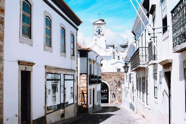 Urlaubsreisen Enge Gasse mit weißen Gebäuden und einem Kirchturm im Hintergrund in der Algarve, Portugal.