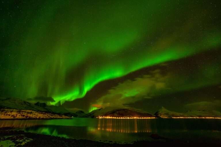 Urlaubsreisen Grüne Polarlichter leuchten über schneebedeckten Bergen und einem ruhigen Gewässer in Norwegen.