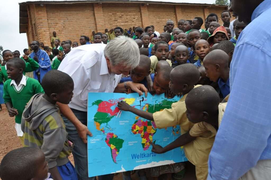 Reiner Meutsch zeigt einer Gruppe von Kindern in Ruanda eine Weltkarte. Die Kinder sind neugierig und zeigen auf verschiedene Länder auf der Karte. Sie befinden sich vor einem Backsteingebäude.