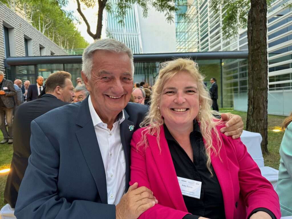 Reiner Meutsch und Anke Budde sitzen lächelnd nebeneinander bei einer Veranstaltung im Freien. Im Hintergrund sind andere Teilnehmer zu sehen.