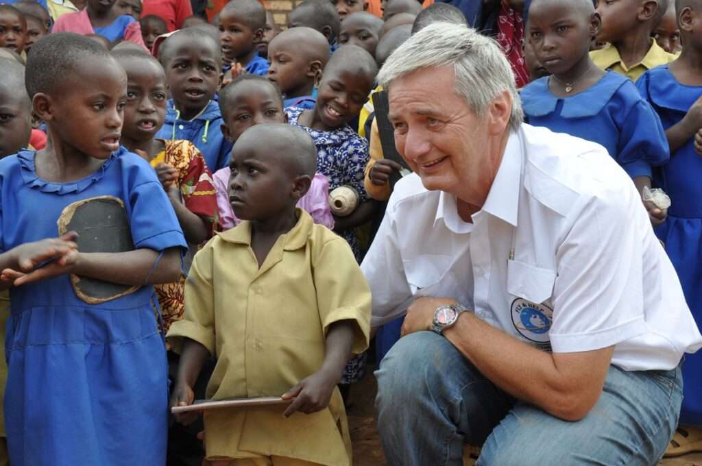 Reiner Meutsch kniet neben einer Gruppe von Kindern in Ruanda, die in bunten Schuluniformen gekleidet sind.