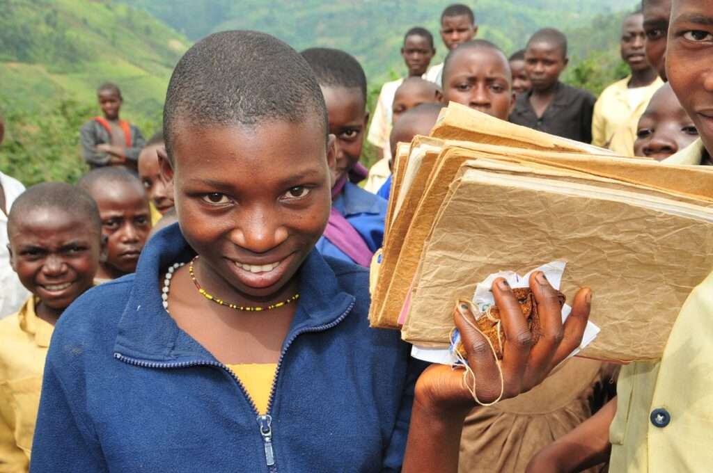 Ein Mädchen lächelt in die Kamera und hält Schulmaterialien in der Hand, während andere Kinder im Hintergrund stehen.