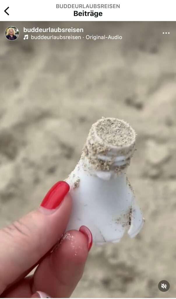 Eine Hand mit roten Fingernägeln hält ein mit Sand bedecktes Stück Plastik am Strand.