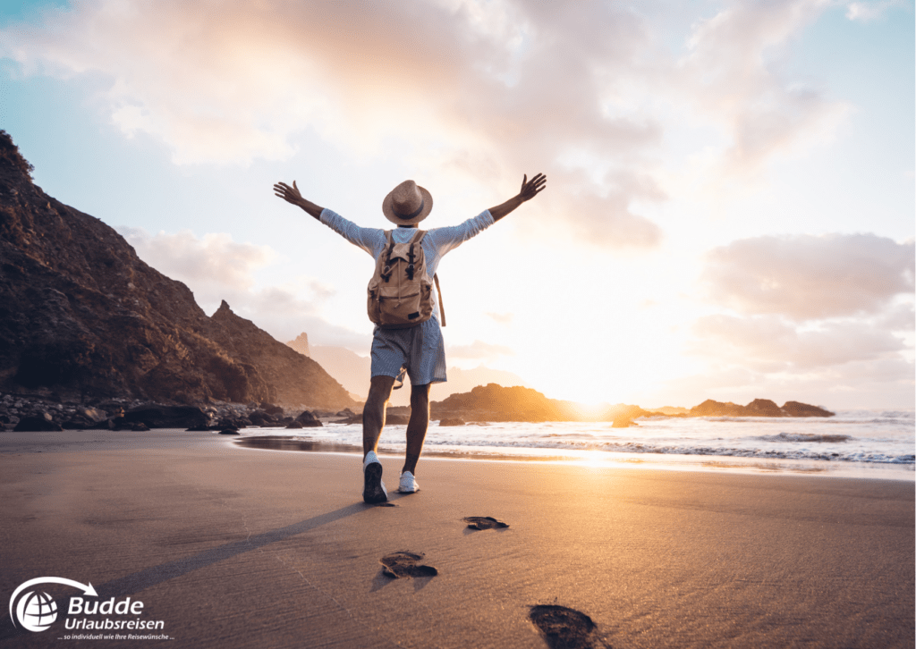 Ein Mann mit Rucksack und Strohhut steht mit ausgestreckten Armen an einem Strand bei Sonnenuntergang und erlebt eine Reise gegen Einsamkeit.