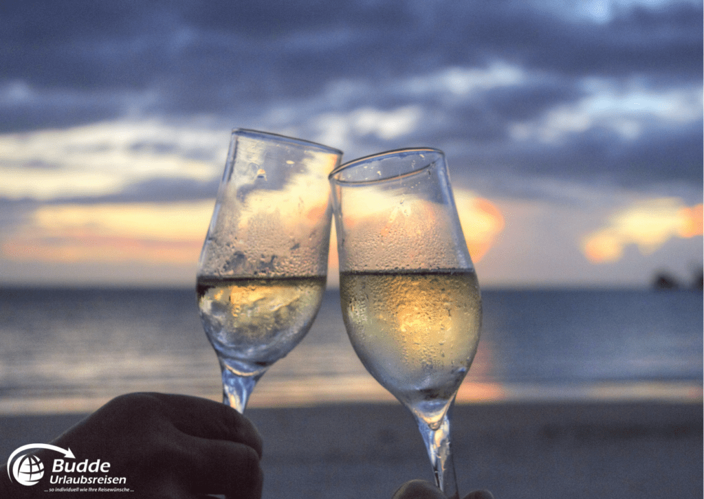 Flitterwochen Saint Martin - Zwei Gläser Champagner stoßen am Strand an, der Sonnenuntergang ist im Hintergrund sichtbar. Reisen nach Saint Martin buchbar bei Budde Urlaubsreisen.
