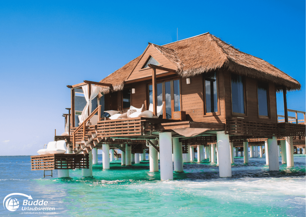 Urlaubsreisen Holzhütte auf Stelzen mit Strohdach, umgeben von klarem Wasser und blauen Himmel.