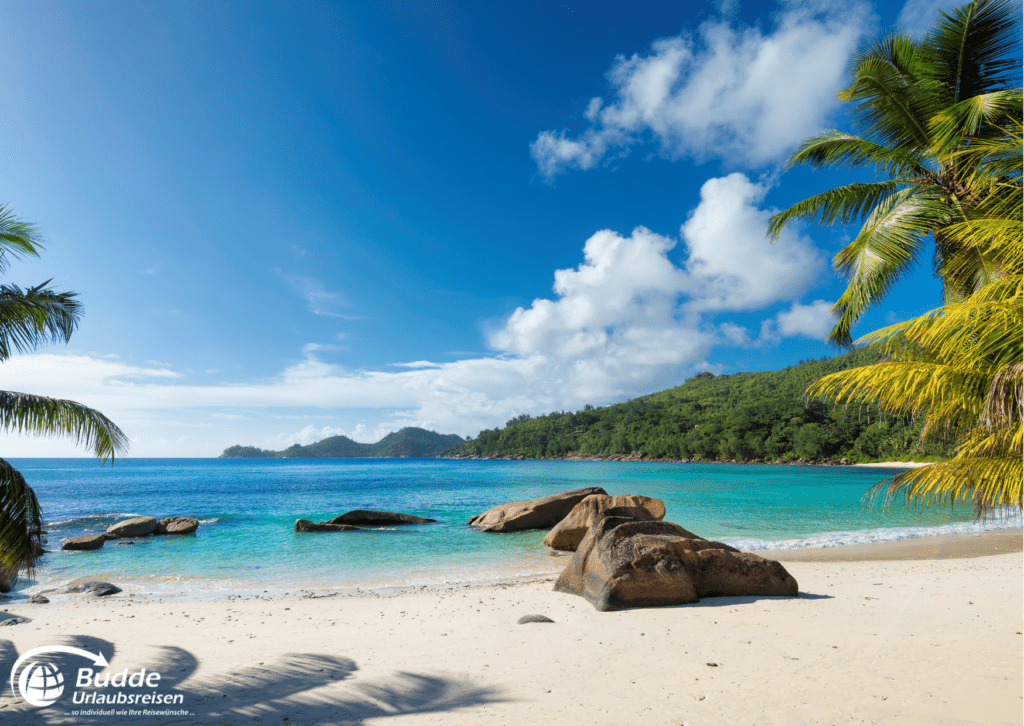 Urlaubsreisen Strand mit feinem Sand, Palmen und Felsen unter einem blauen Himmel mit Wolken.