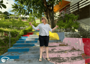 Reiseerfahrungen Saint Martin: Eine Frau steht vor bunten Treppenstufen und posiert für ein Foto, angeboten von Budde Urlaubsreisen.