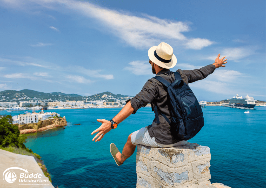 Ein Mann sitzt auf einer Mauer mit Blick auf das Meer und eine Stadt im Hintergrund und erlebt eine Reise gegen Einsamkeit.