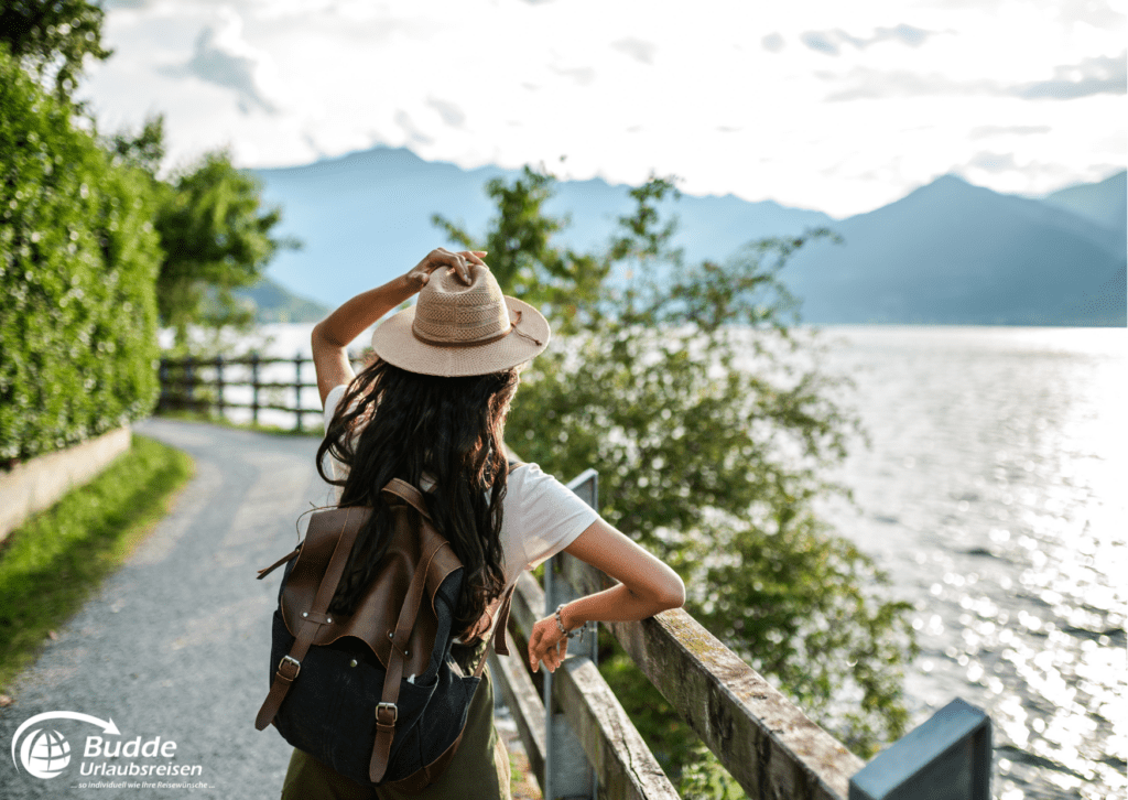 Eine Frau mit Hut und Rucksack blickt auf einen See mit Bergen im Hintergrund, eine perfekte Reise gegen Einsamkeit.