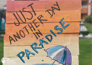 Ägypten Urlaub: Holztafel mit dem Schriftzug 'Just Another Day in Paradise' und einem Regenschirm-Motiv. - buchbar im Reisebüro Budde Urlaubseisen