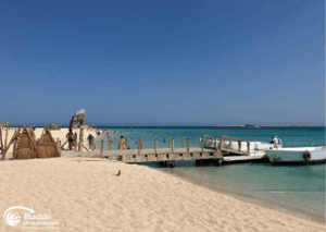 Ägypten Urlaub: Holzsteg, der zu einem Strand in Marsa Alam führt, mit Booten im Wasser und Sonnenschein. - buchbar im Reisebüro Budde Urlaubseisen