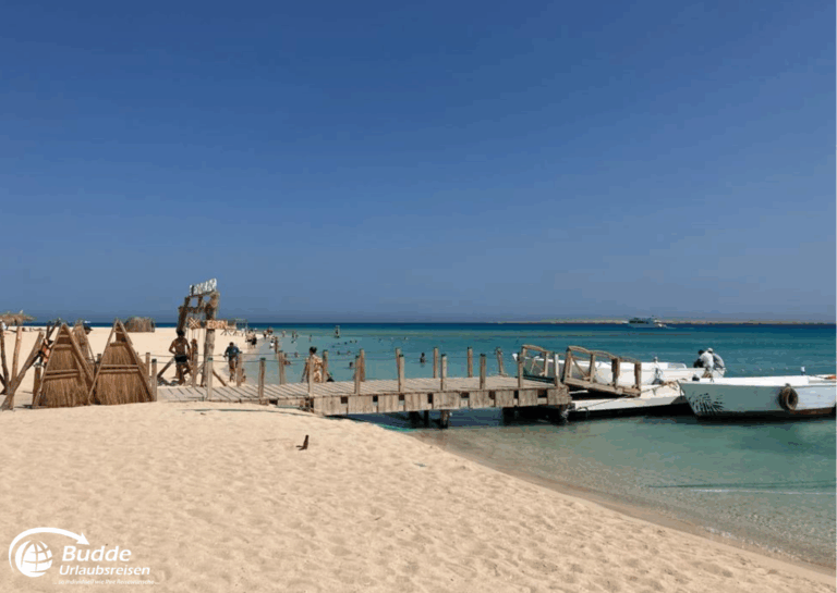 Ägypten Urlaub: Holzsteg, der zu einem Strand in Marsa Alam führt, mit Booten im Wasser und Sonnenschein. - buchbar im Reisebüro Budde Urlaubseisen