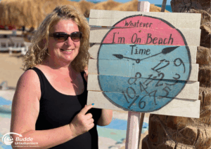Ägypten Urlaub: Reiseexpertin Anke Budde steht neben einem Schild mit der Aufschrift 'I'm On Beach Time' und einer bunten Uhr am Strand. - buchbar im Reisebüro Budde Urlaubseisen