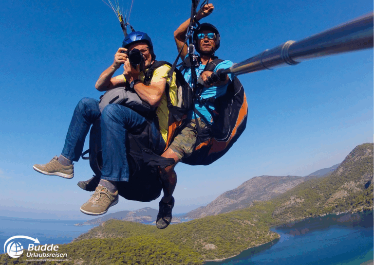 Diamir Erlebnisreisen Zwei Personen beim Paragliding über eine Küstenlandschaft mit Bergen und Wasser im Hintergrund. - buchbar im Reisebüro Budde Urlaubseisen