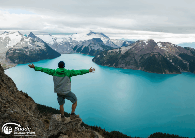 Diamir Erlebnisreisen Mann steht mit ausgebreiteten Armen auf einem Felsen mit Blick auf einen blauen See und Berge. - buchbar im Reisebüro Budde Urlaubseisen
