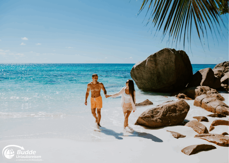 Diamir Erlebnisreisen Ein Paar, das am Strand der Seychellen entlanggeht, mit Wasser und Felsen im Hintergrund. - buchbar im Reisebüro Budde Urlaubseisen