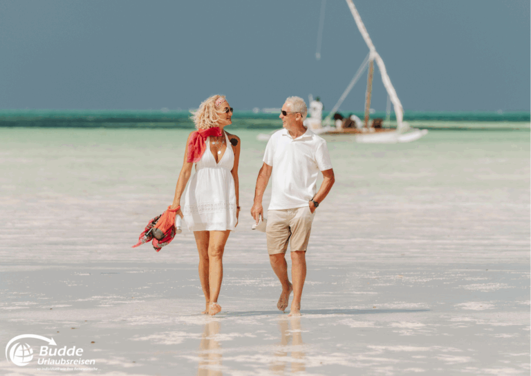 Langzeiturlaub buchen: Ein Paar in leichter Kleidung geht barfuß am Strand entlang, mit einem Segelboot im Hintergrund. - buchbar im Reisebüro Budde Urlaubseisen
