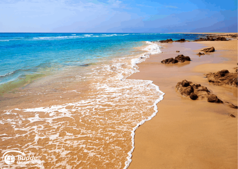 Langzeitreise buchen: Sandstrand mit sanften Wellen und Felsen am Ufer unter blauem Himmel. - buchbar im Reisebüro Budde Urlaubseisen