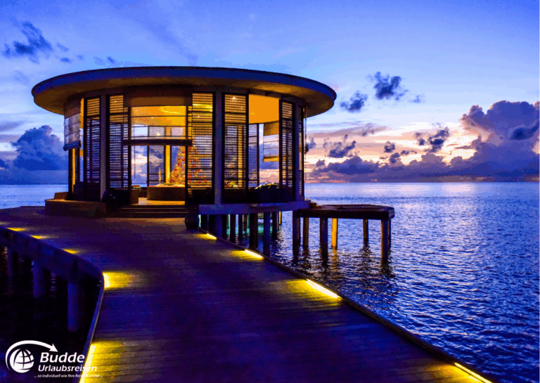 Überwasser-Bungalow mit Holzsteg und beleuchtetem Eingang bei Sonnenuntergang auf den Malediven. - Dein Luxusurlaub buchbar im Reisebüro Budde Urlaubseisen