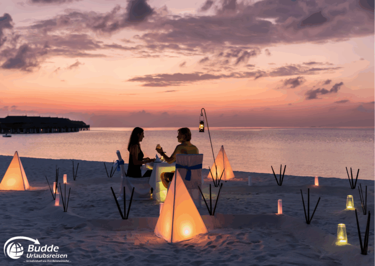 Zwei Personen sitzen an einem Tisch am Strand bei Sonnenuntergang, umgeben von leuchtenden Laternen und Zeltstrukturen. - buchbar im Reisebüro Budde Urlaubseisen