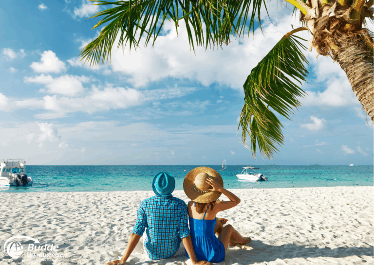 Ein Paar sitzt am Strand der Malediven mit Blick auf das Meer und Palmblätter im Vordergrund. - buchbar im Reisebüro Budde Urlaubseisen