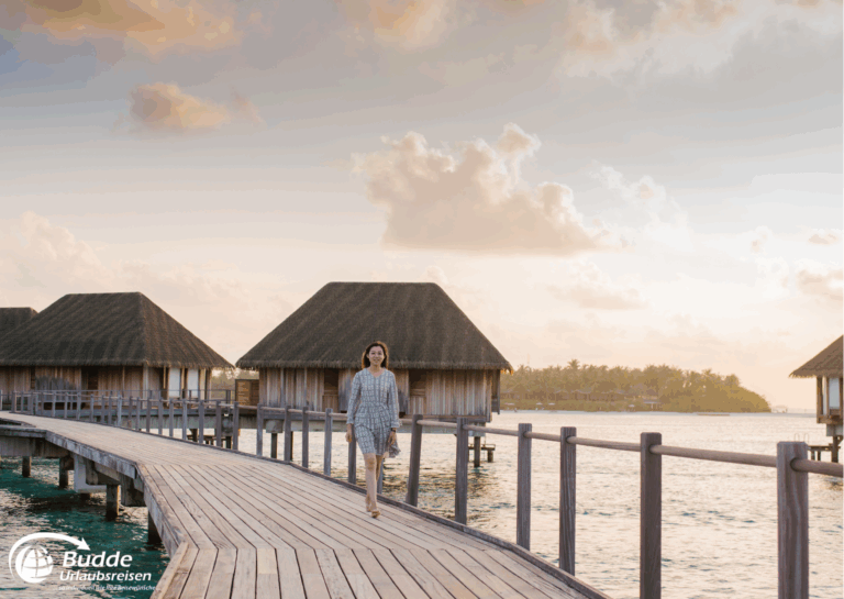 Eine Frau geht auf einem Holzsteg zu den Wasserbungalows in den Malediven bei Sonnenuntergang. - buchbar im Reisebüro Budde Urlaubseisen
