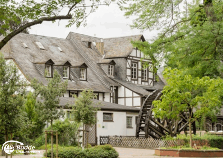 Urlaub an der Nahe: Historisches Gebäude mit Fachwerk und Wassermühle im Vordergrund, umgeben von Bäumen und Grünflächen. - buchbar im Reisebüro Budde Urlaubseisen