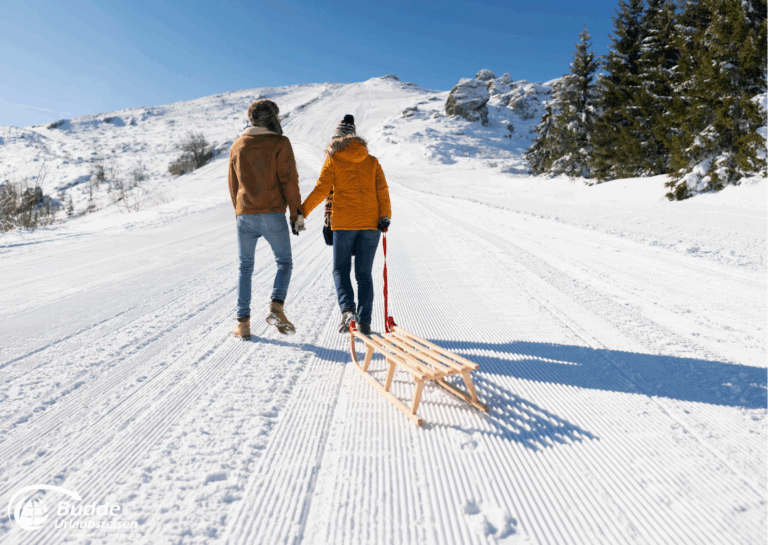 Romantikurlaub zu Zweit: Ein Paar hält sich an den Händen und zieht einen Holzschlitten auf einer verschneiten Piste. - buchbar im Reisebüro Budde Urlaubseisen