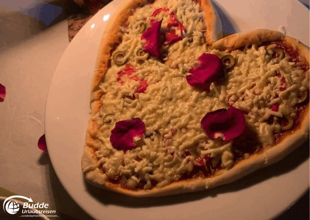 Praana Ayurvedic Boutique Hotel: Herzförmige Pizza mit Käse, Tomatensauce und dekorativen Blütenblättern auf einem Teller. - buchbar im Reisebüro Budde Urlaubseisen