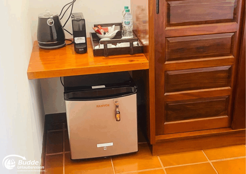 Kompakter Kühlschrank und Kaffeezubereiter auf einem Holzregal in einem Zimmer. - buchbar im Reisebüro Budde Urlaubseisen
