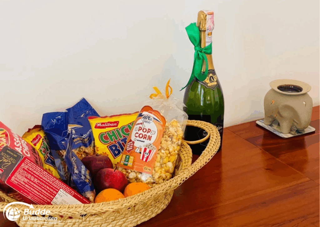 Ein Korb mit Snacks und Obst neben einer Champagnerflasche auf einem Tisch. - buchbar im Reisebüro Budde Urlaubseisen