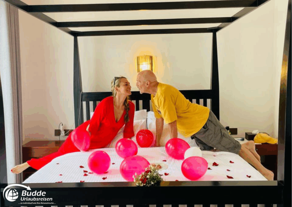 Ein Paar sitzt auf einem Bett mit roten und rosa Luftballons sowie Blütenblättern im Schlafzimmer. - buchbar im Reisebüro Budde Urlaubseisen