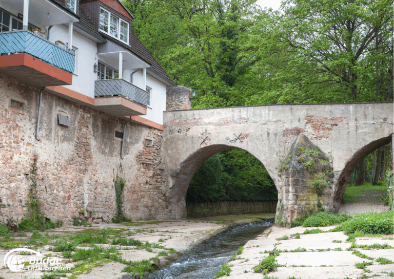 Urlaub an der Nahe: Steinbrücke mit zwei Bögen über einen kleinen Fluss in Bad Kreuznach, umgeben von Bäumen. - buchbar im Reisebüro Budde Urlaubseisen