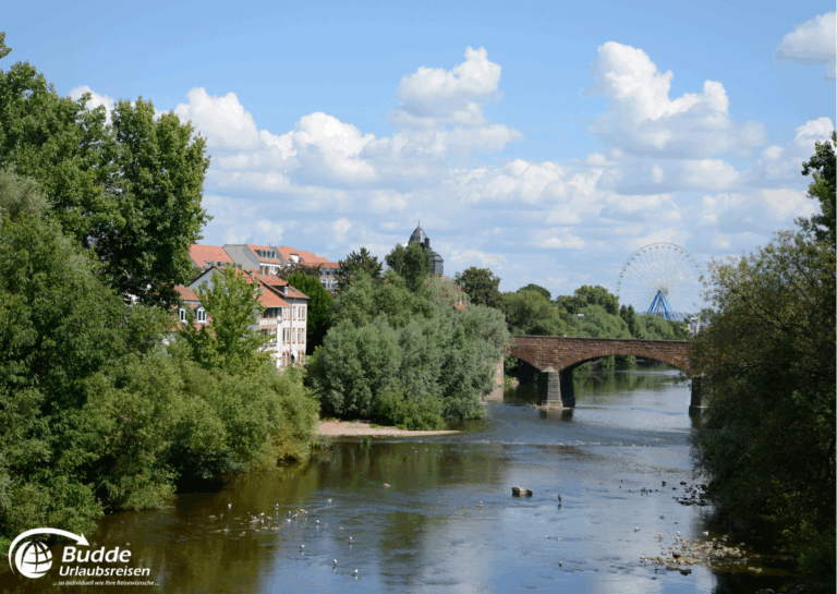 Urlaub an der Nahe: Brücke über einen Fluss in Bad Kreuznach mit Bäumen und einem Riesenrad im Hintergrund. - buchbar im Reisebüro Budde Urlaubseisen