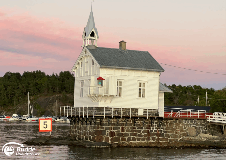 Norwegen Urlaub: Weißes Holzgebäude mit spitzem Dach und Turm, umgeben von Wasser und Booten. Norwegen Urlaub: Weißes Holzgebäude mit spitzem Dach und Turm, umgeben von Wasser und Booten.