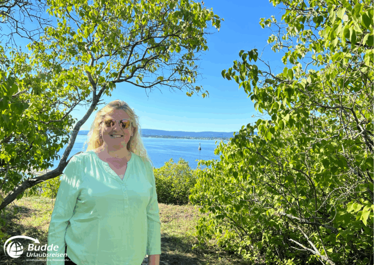 Norwegen Urlaub: Eine Frau mit blonden Haaren und Sonnenbrille steht vor einem Gewässer, umgeben von Bäumen. Norwegen Urlaub: Eine Frau mit blonden Haaren und Sonnenbrille steht vor einem Gewässer, umgeben von Bäumen.
