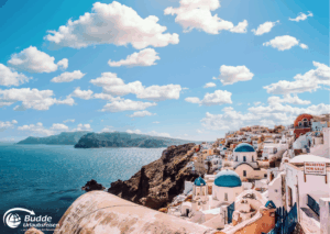 Strandurlaub Griechenland Insel Mykonos Panoramaansicht der Küste von Mykonos mit weißen Gebäuden und blauen Kuppeln unter einem bewölkten Himmel.