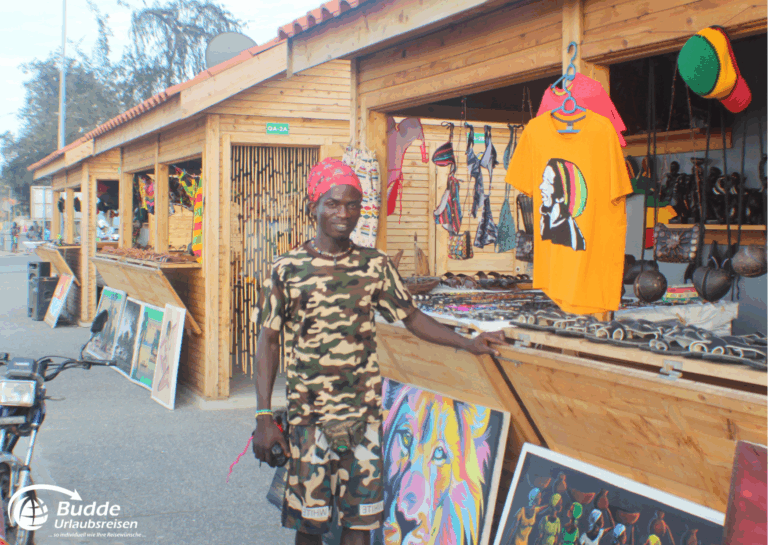 Mann in Camouflage-Kleidung steht vor einem Marktstand mit verschiedenen Waren in Angola. - Urlaub in Angola buchen