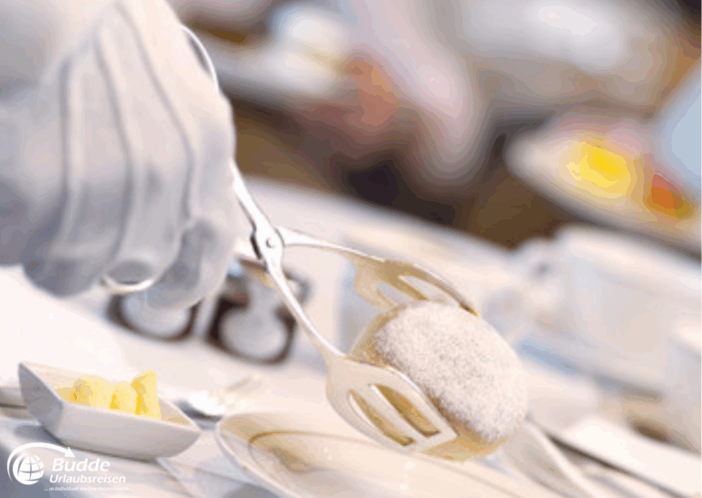 Weltreise Queen Mary 2: Ein Kellner in Handschuhen hebt ein zuckerbestäubtes Gebäck mit einer Zange auf einem Tisch an.