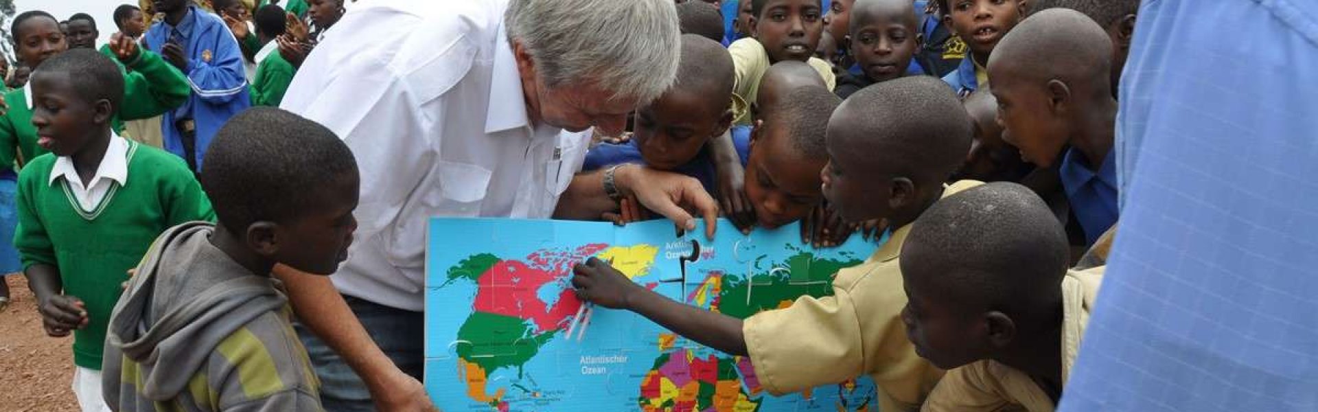 Reiner Meutsch zeigt einer Gruppe von Kindern in Ruanda eine Weltkarte. Die Kinder sind neugierig und zeigen auf verschiedene Länder auf der Karte. Sie befinden sich vor einem Backsteingebäude.